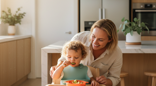 Toddler self-feeding with supportive parent in warm kitchen